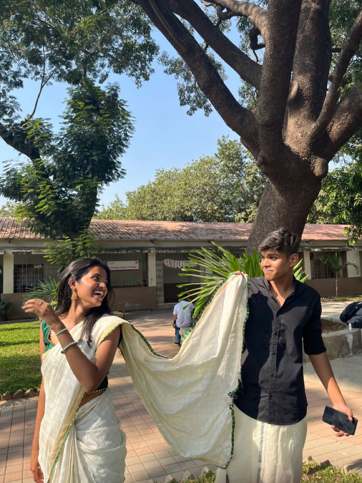 two people standing under a tree in front of a house and one is holding a white sari