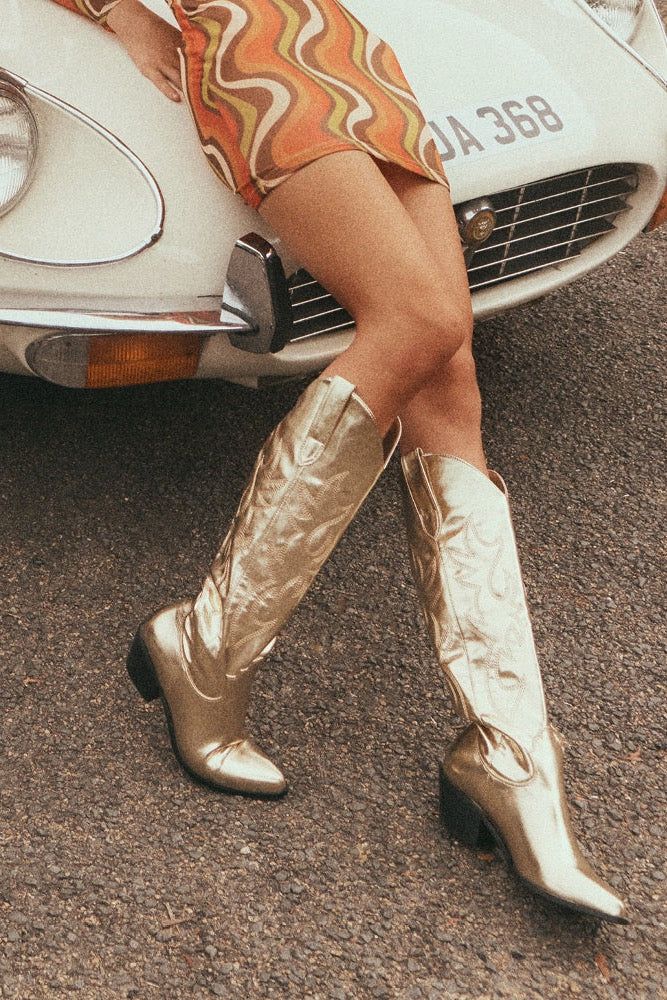 Stand out from the crowd with this statement long boot! The Urson by Billini is a long, western inspired boot. It features a low stacked block heel, wide fit long boot body, western inspired embroidery, an almond shape toe, and an eye-catching metallic silver finish. We guarantee these will quickly become your favorite way to jazz up any look! Run true to size ~ if in between sizes then we recommend sizing up Heel height is 5.5 cm Oversized side pull tabs for a pull on design Synthetic upper, li Gold Metallic Cowboy Boots, Billini Cowboy Boots, Wide Calf Cowgirl Boots, Knee High Cowgirl Boots, High Cowgirl Boots, Boot Stand, Knee High Leather Boots, Gold Heels, Long Boots