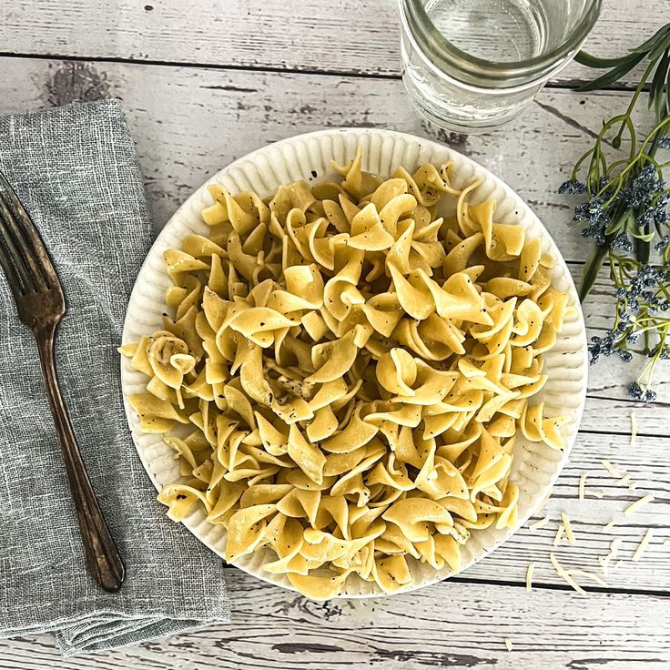 a white plate topped with pasta next to a fork and glass filled with water on top of a wooden table