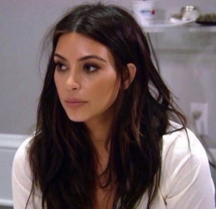 a woman with long dark hair sitting in front of a bathroom sink and looking at the camera