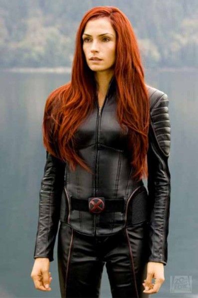 a woman with long red hair standing in front of a body of water wearing black leather