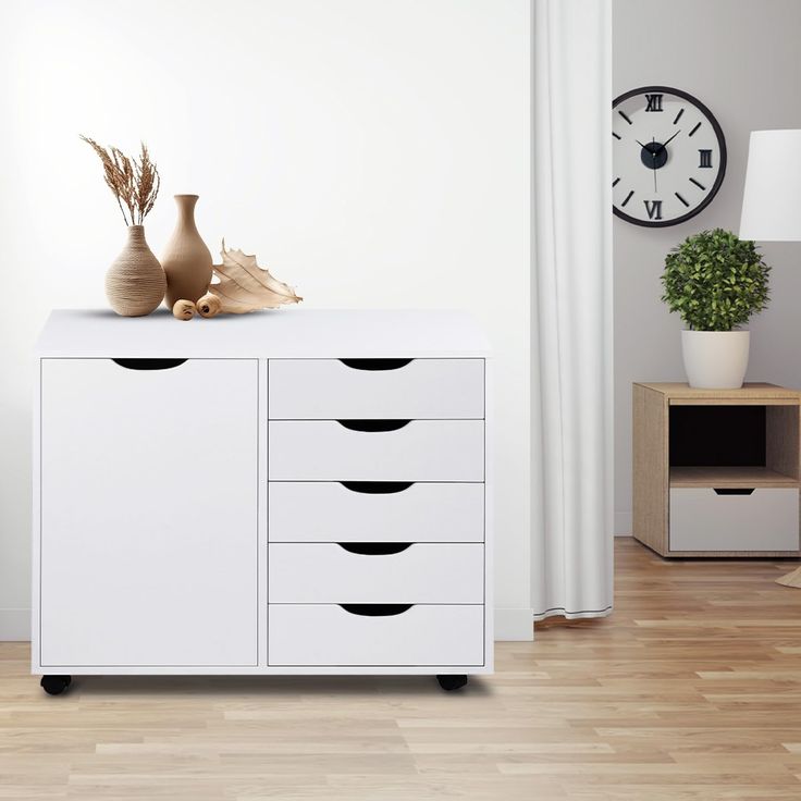 a white cabinet with five drawers in front of a wall clock and vases on top