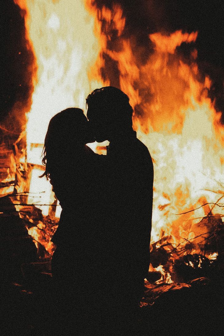 two people standing in front of a fire with their arms around each other and kissing