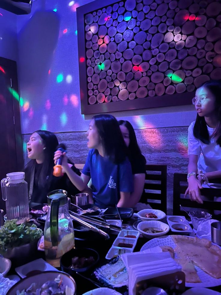 three women sitting at a table with food in front of them and lights on the wall behind them