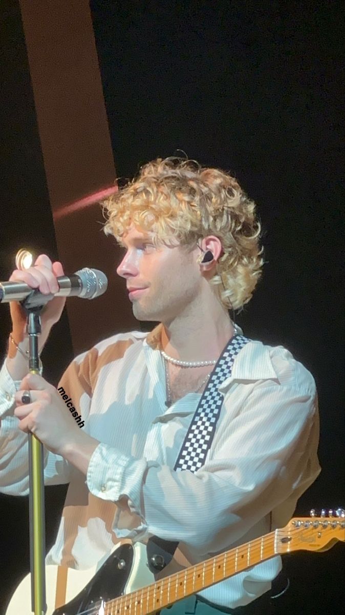 a man with blonde hair is singing into a microphone and holding a guitar in front of him