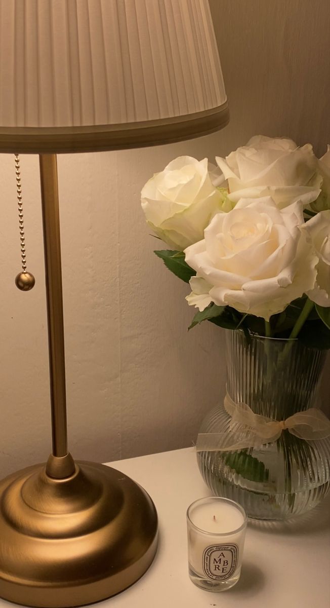 white roses in a vase on a table next to a candle and a lamp shade