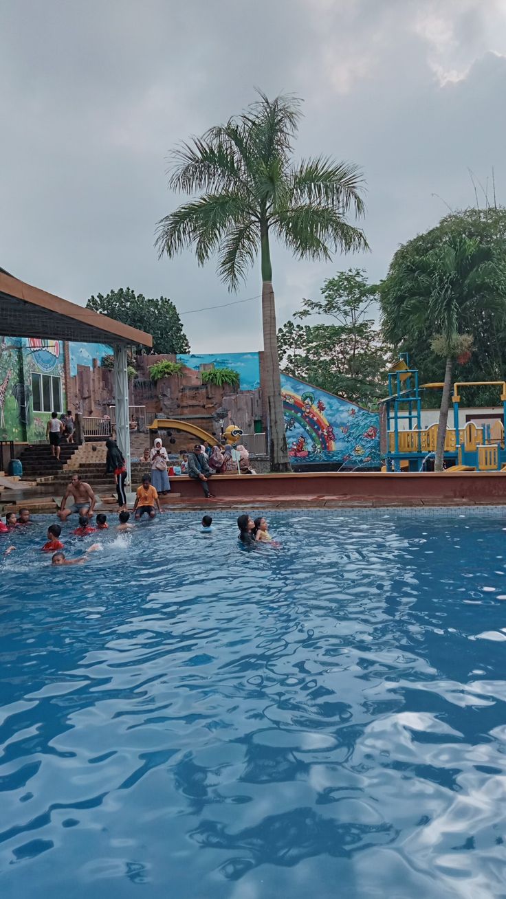 people are swimming in the pool with blue water and palm trees behind them on a cloudy day