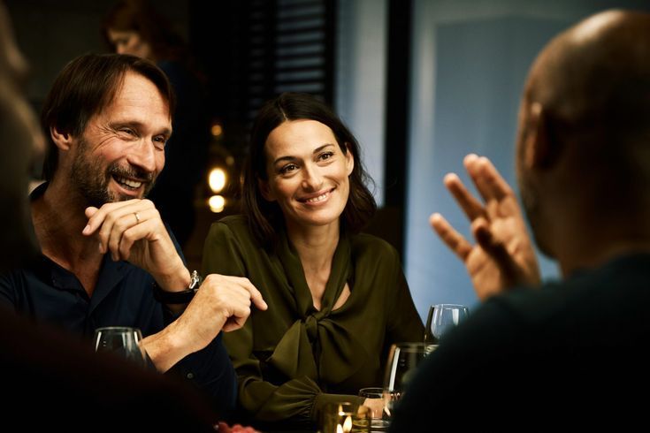 a man and woman sitting at a table talking to each other with wine glasses in front of them