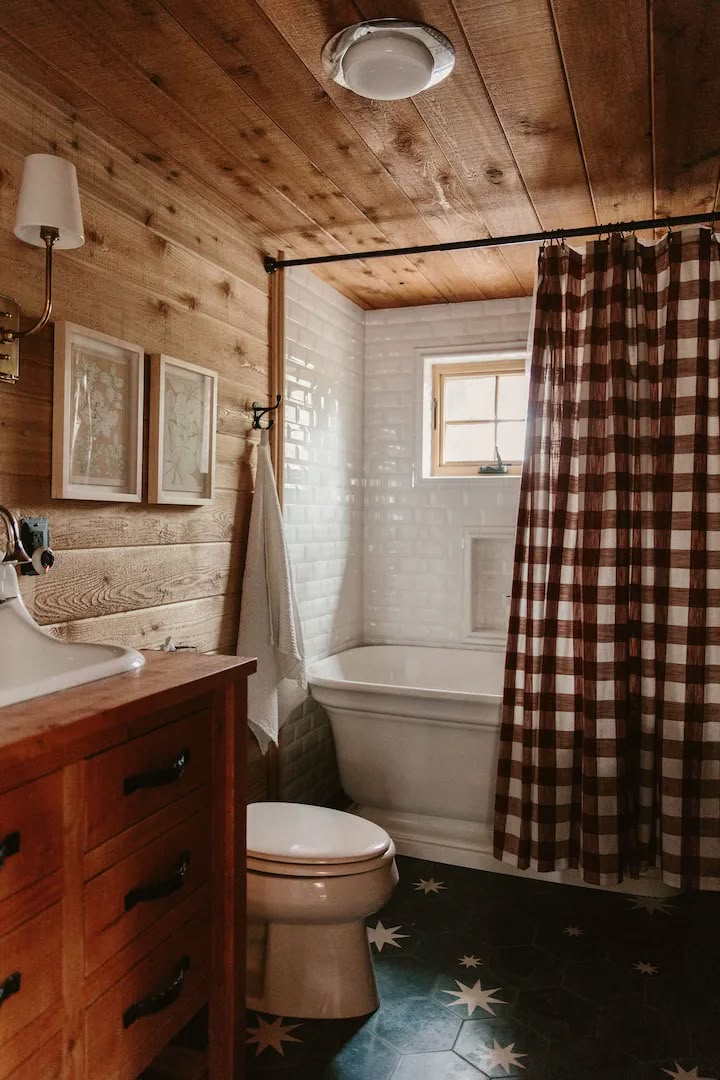 a white toilet sitting next to a bath tub under a bathroom window covered in checkered curtains