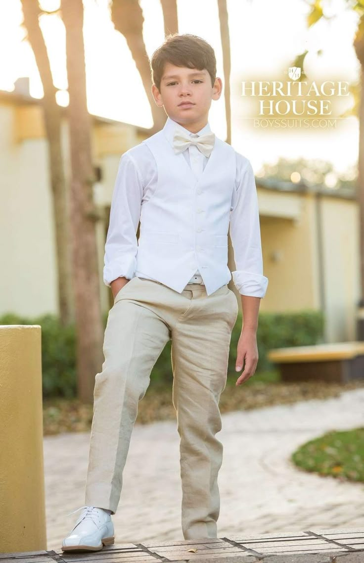 a young boy in a white shirt and bow tie standing on a brick walkway with palm trees behind him