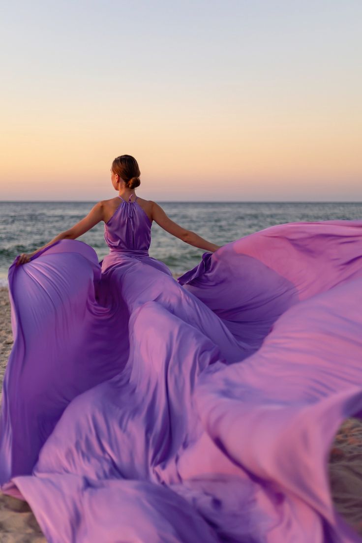 a woman in a long purple dress is standing on the beach with her back to the camera