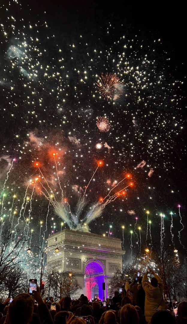 fireworks are lit up in the night sky above a monument and trees as people watch