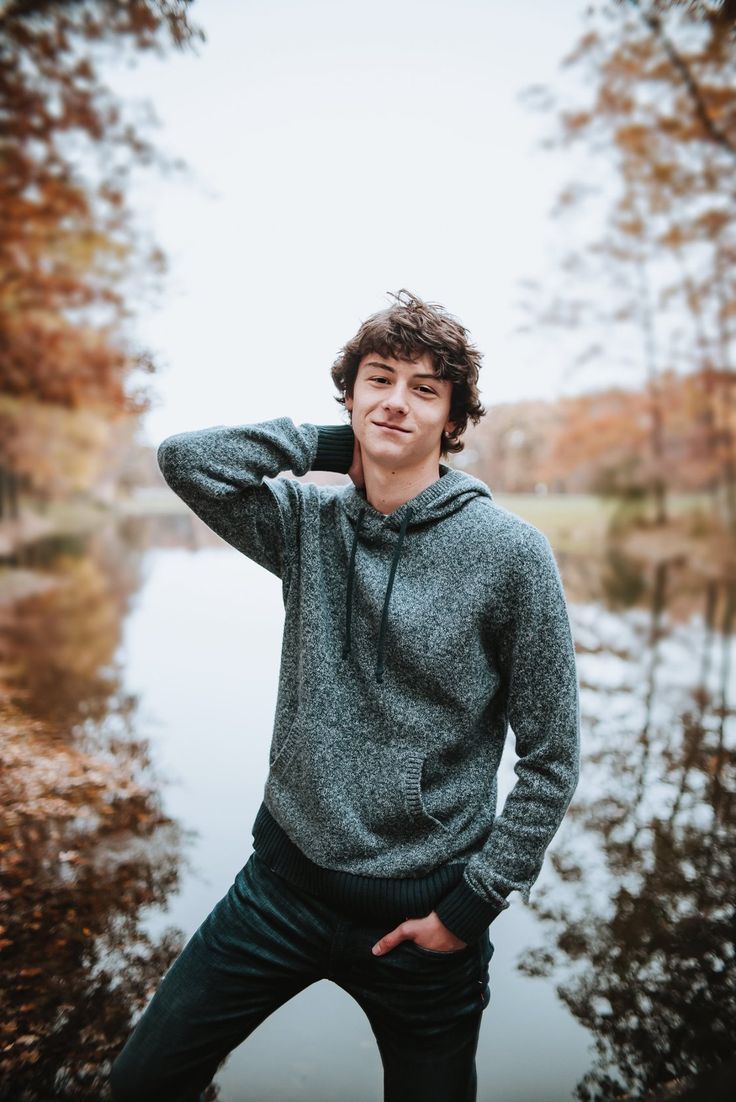 a young man poses for a photo in front of a pond with trees and leaves