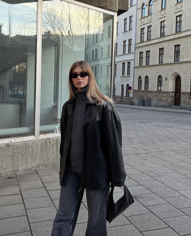 a woman in black jacket and jeans standing on sidewalk next to building with large windows