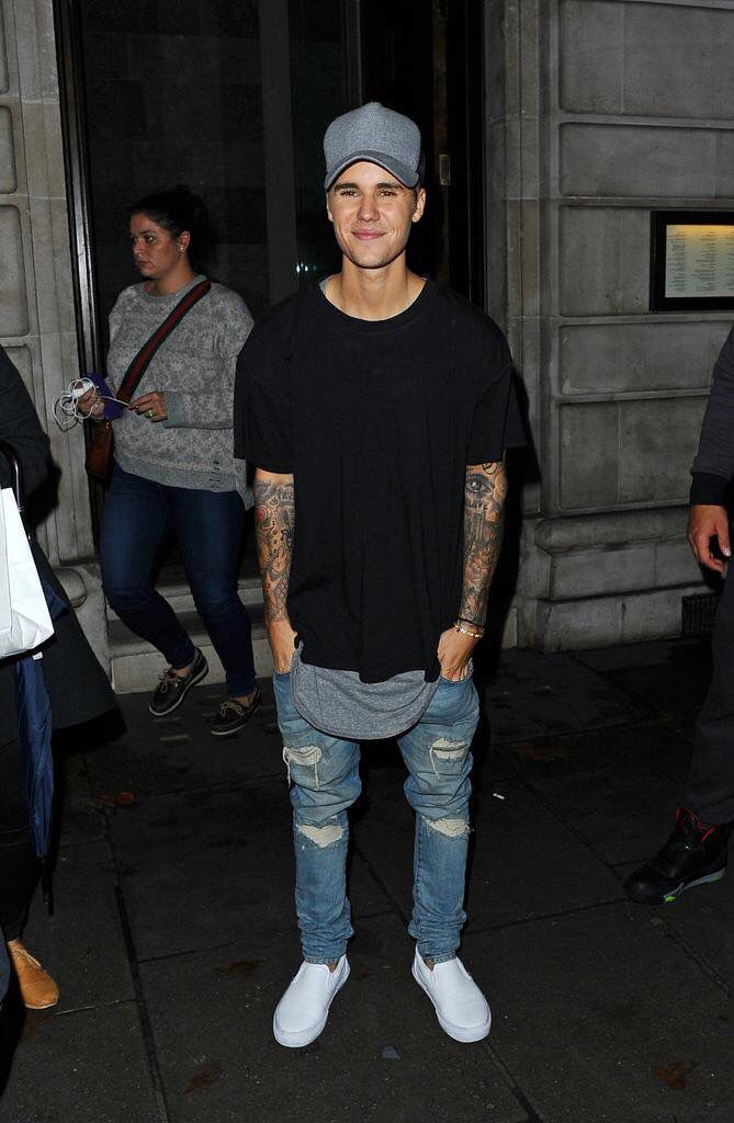 a man standing in front of a building wearing ripped jeans and a baseball cap with tattoos on his arm