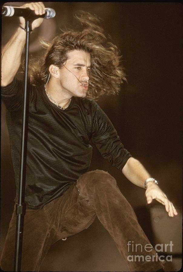 a male in a black shirt is on stage with a microphone and hair blowing in the wind