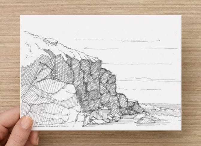 a hand holding up a card with a drawing of rocks on the water and mountains in the background