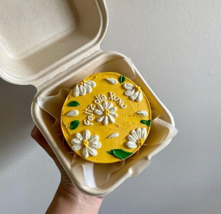 a hand holding a yellow and white decorated cake in a foam container with the lid open