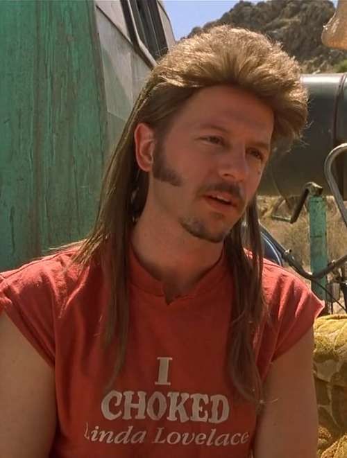 a man with long hair sitting down in front of a truck and looking at the camera