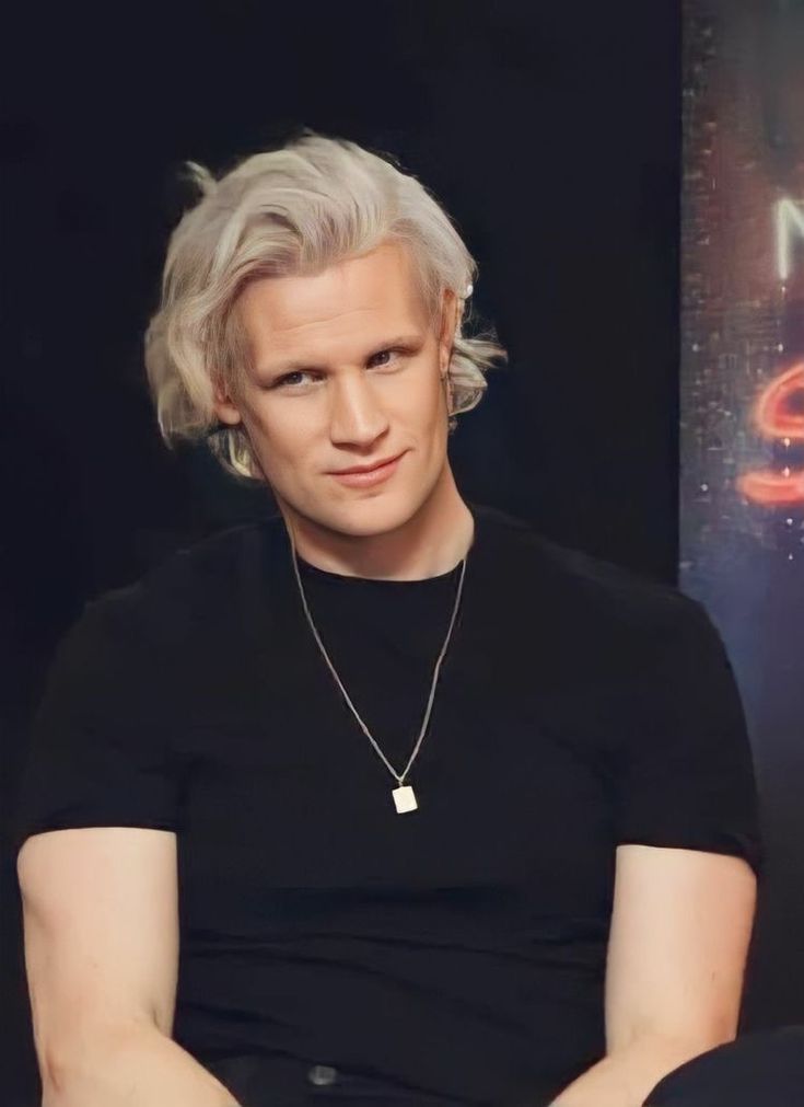 a man with grey hair sitting in front of a black wall and wearing a black t - shirt