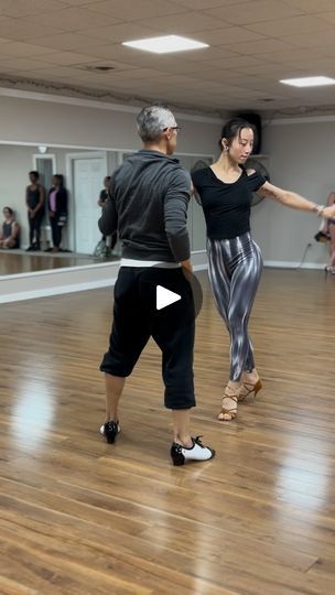 two people dancing on a wooden floor in an empty room with other people standing around