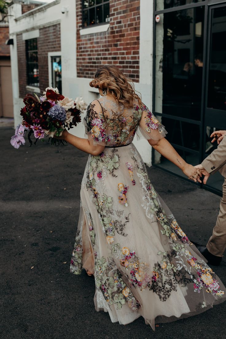 a bride and groom holding hands walking down the street with flowers in their hand,