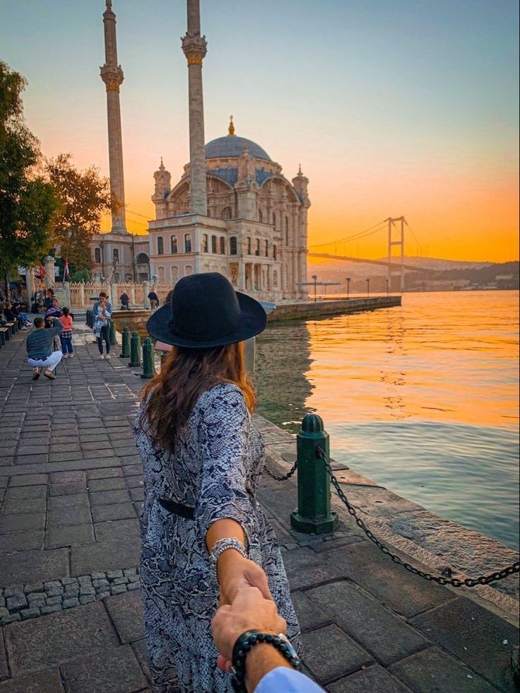 a woman holding the hand of a man in front of a body of water at sunset