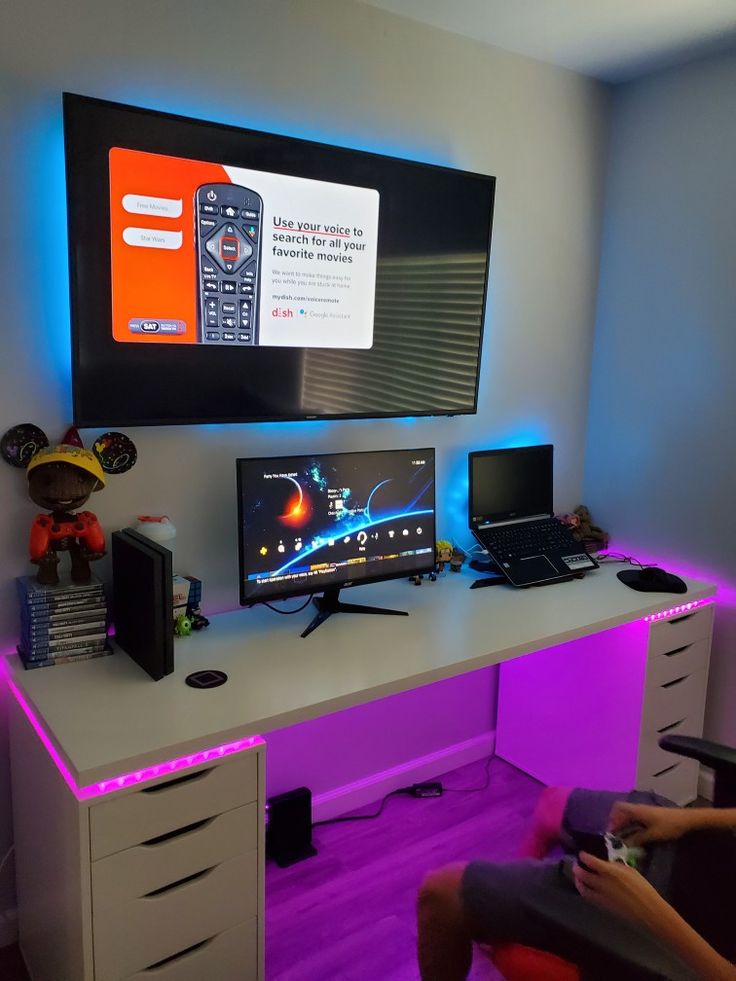 a computer desk with two monitors and a laptop on it, in front of a flat screen tv