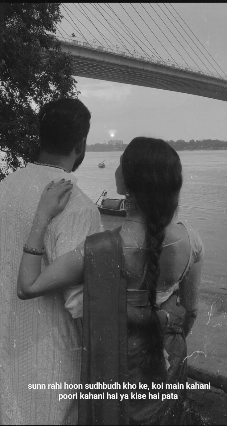 a man and woman standing next to each other near the water with a bridge in the background