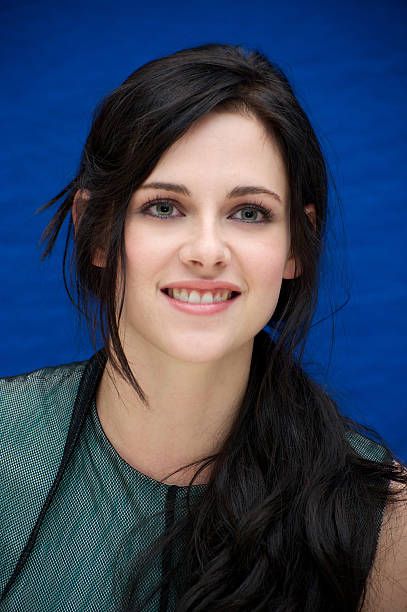 a woman with long black hair wearing a green top and smiling at the camera in front of a blue background