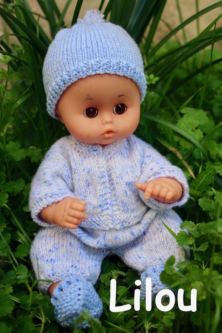 a little baby doll sitting in the grass wearing a blue knitted outfit and hat