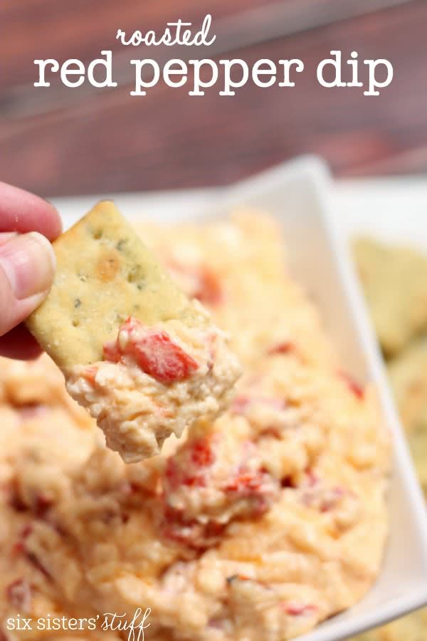 a hand holding a cracker over a bowl of red pepper dip