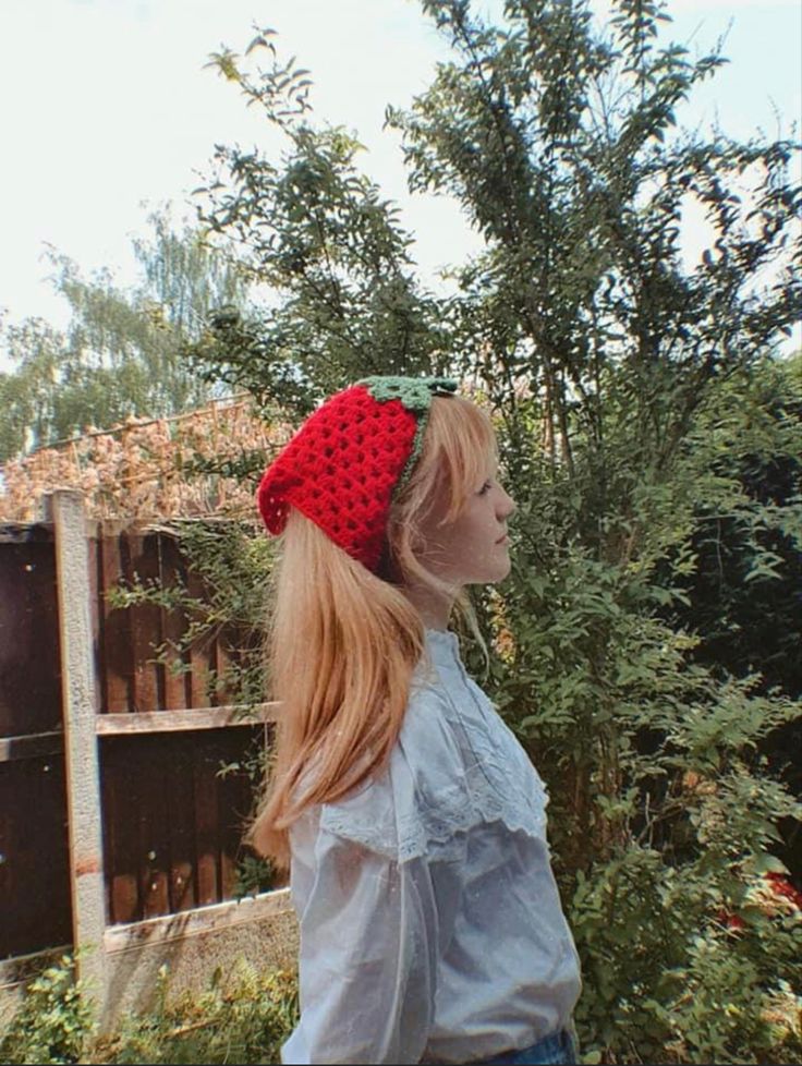 a woman wearing a red knitted hat standing in front of some bushes and trees