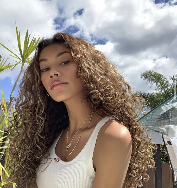 a beautiful young woman with long curly hair standing in front of a fence and palm tree