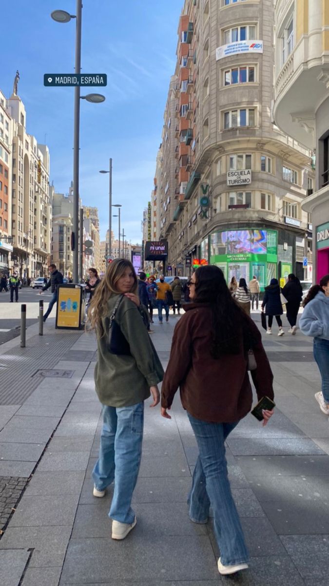 two women walking down the street in front of tall buildings and people on the sidewalk