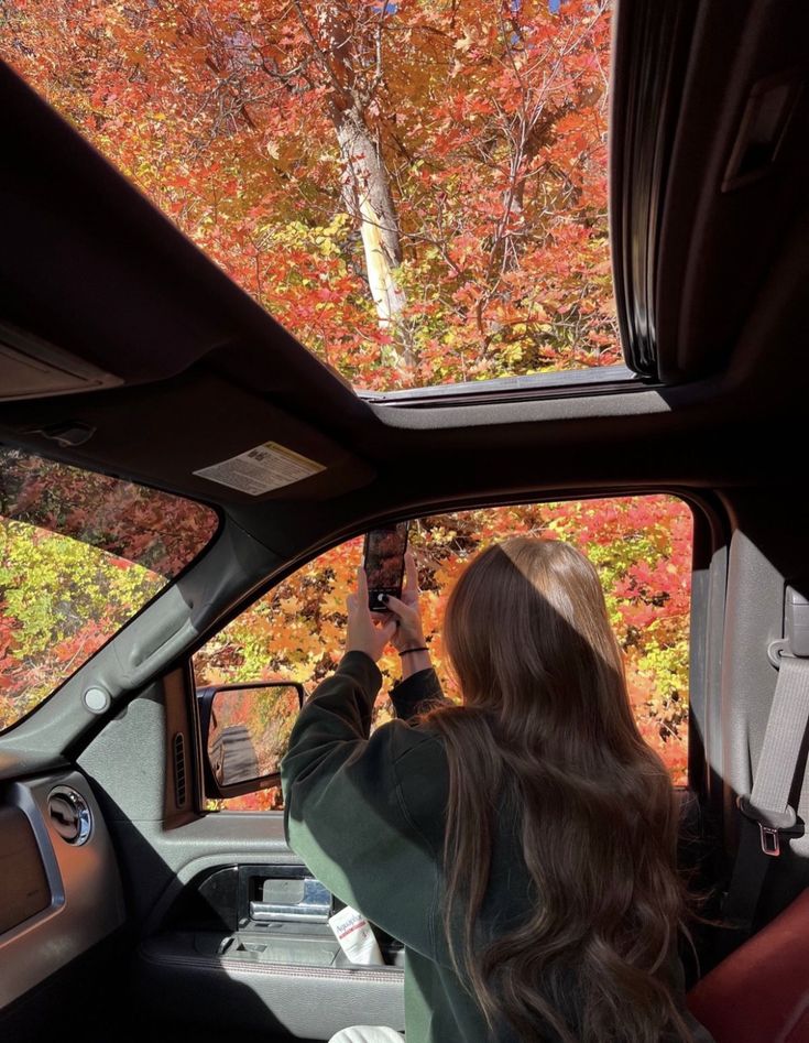 a woman taking a photo in the side mirror of a car with fall foliage behind her