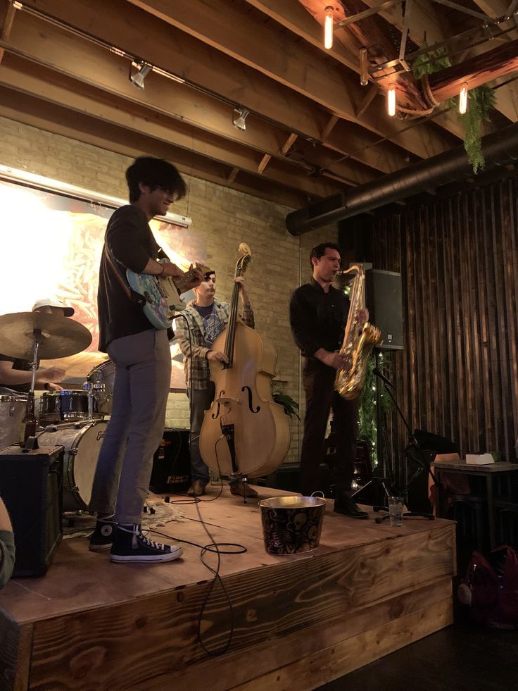 three men are playing instruments on stage in a room with wood floors and exposed ceilings