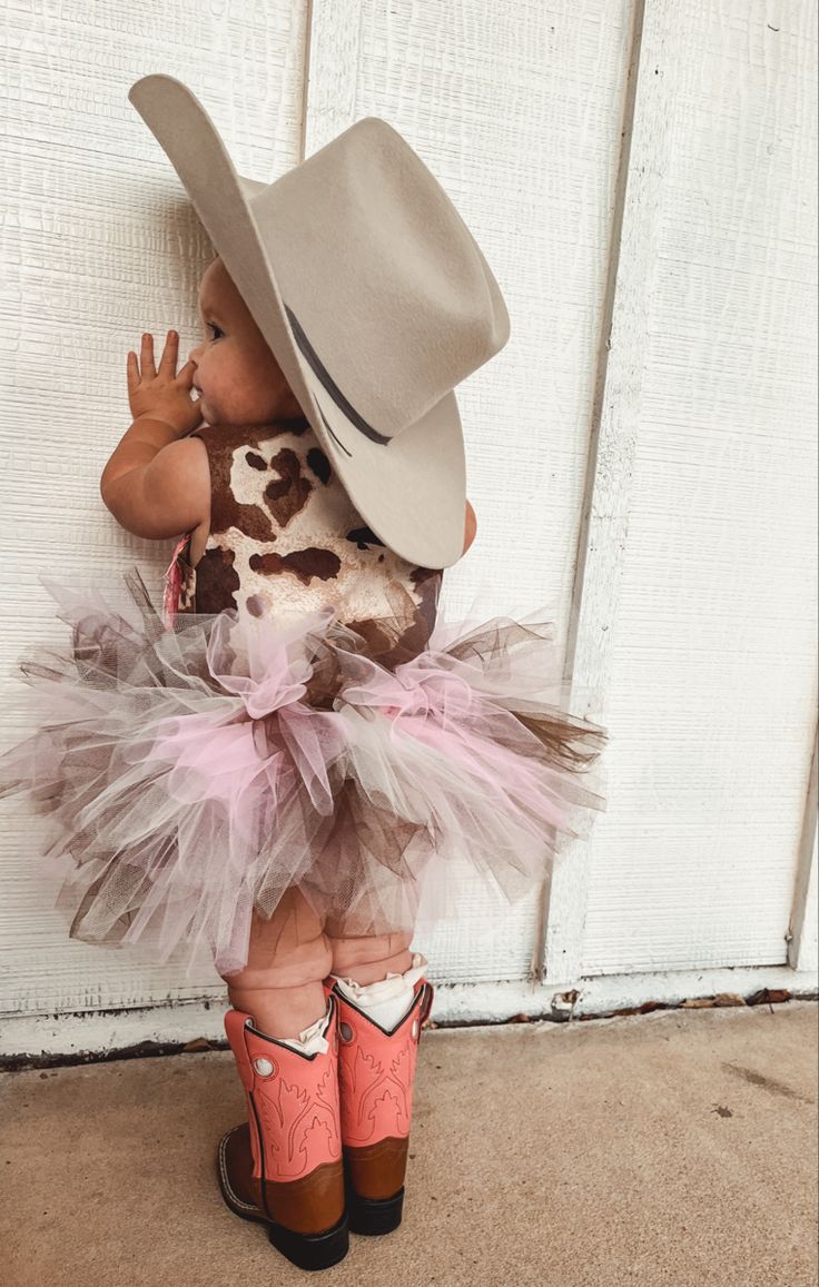 a small child wearing a cowboy hat and pink tutu skirt, standing in front of a white door