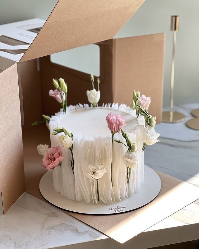 a white cake with pink and white flowers sitting on top of a table next to a cardboard box