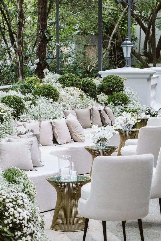 a bunch of white couches and chairs in a room filled with trees, bushes and flowers