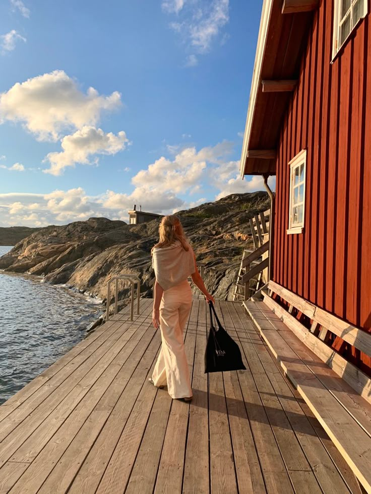 a woman walking down a wooden pier next to a red building and body of water