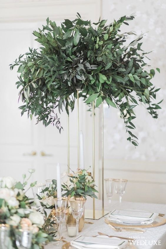 the table is set with white and gold place settings, greenery in tall vases