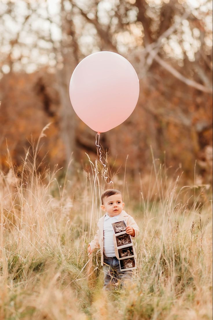 a young boy holding a pink balloon while standing in tall grass