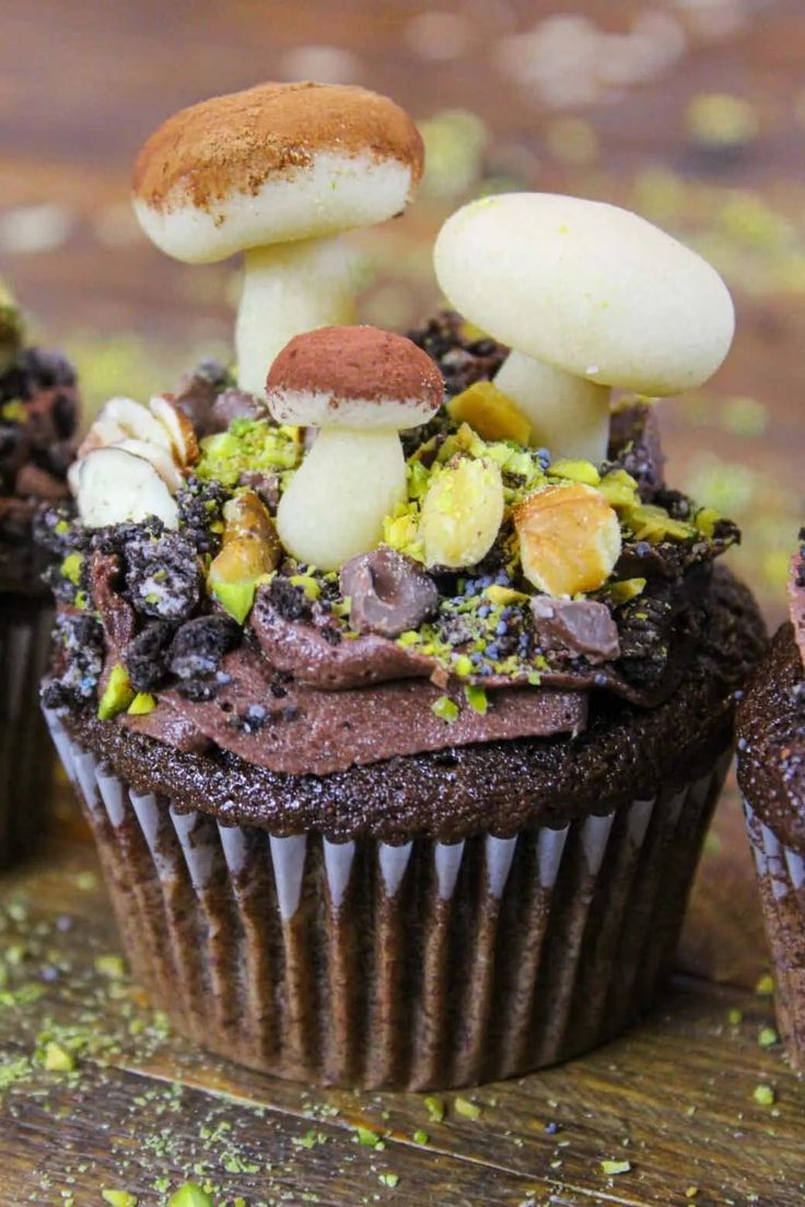 three chocolate cupcakes with mushrooms and other toppings on top, sitting on a wooden table