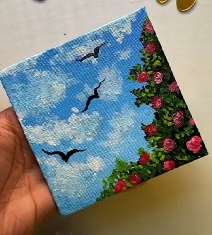 a hand holding up a small painting with birds flying above it and roses in the foreground