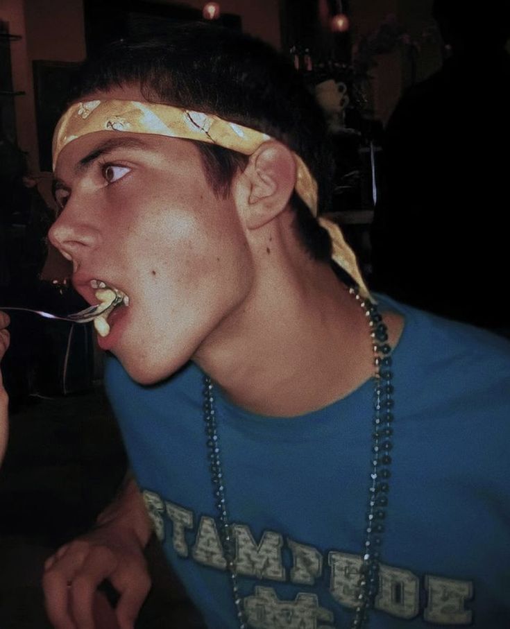 a young man eating food with a spoon in his mouth while wearing a bandana