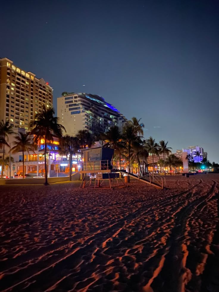 an empty beach at night with palm trees and buildings in the backgrouds