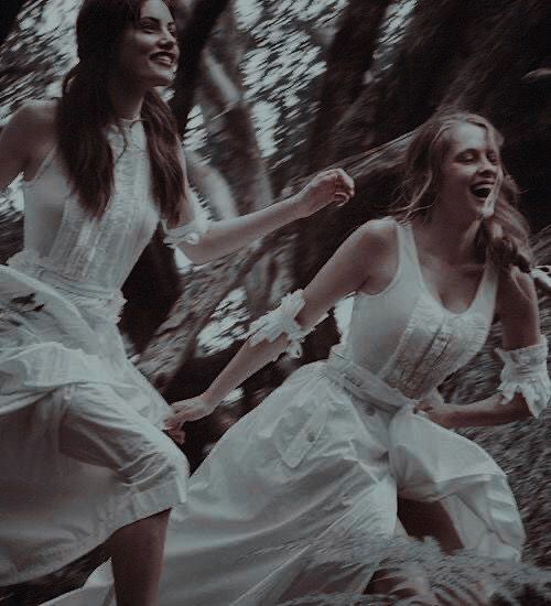 two women in white dresses are running through the woods with their hands together and laughing