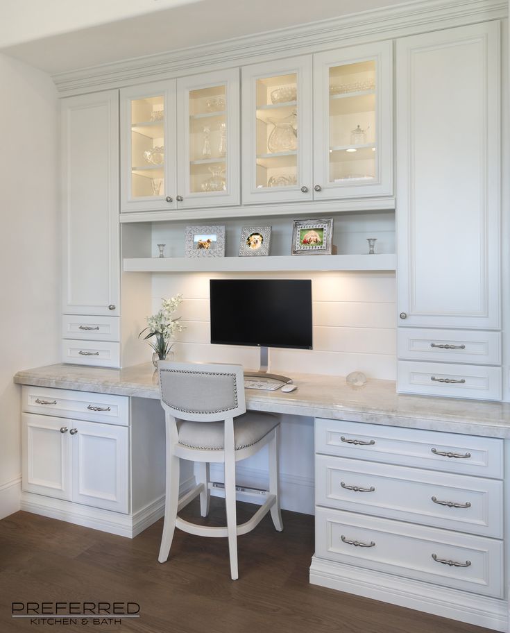 a desk with a computer on top of it in front of white cabinets and drawers