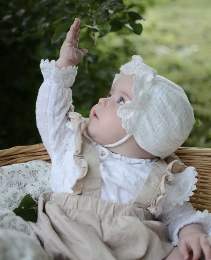 a baby in a white dress and bonnet sitting on a wicker basket